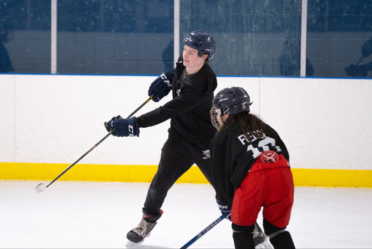 Two hockey players on an ice rink, one in black jersey and the other in red jersey, both wearing helmets and holding hockey sticks.